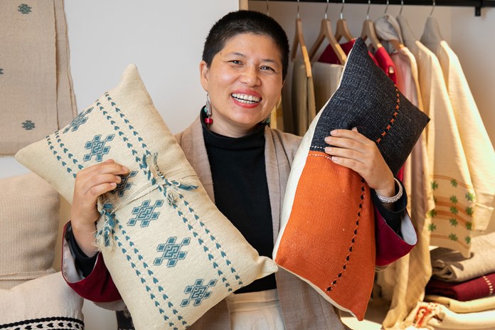 The image shows a person holding two decorative pillows against a backdrop of a clothing rack. One pillow is cream with blue embroidery, the other is half black, half orange with stitched details. The individual is dressed in a beige jacket over a burgundy top, and an earring is visible. The setting suggests a retail store or cozy interior space.
