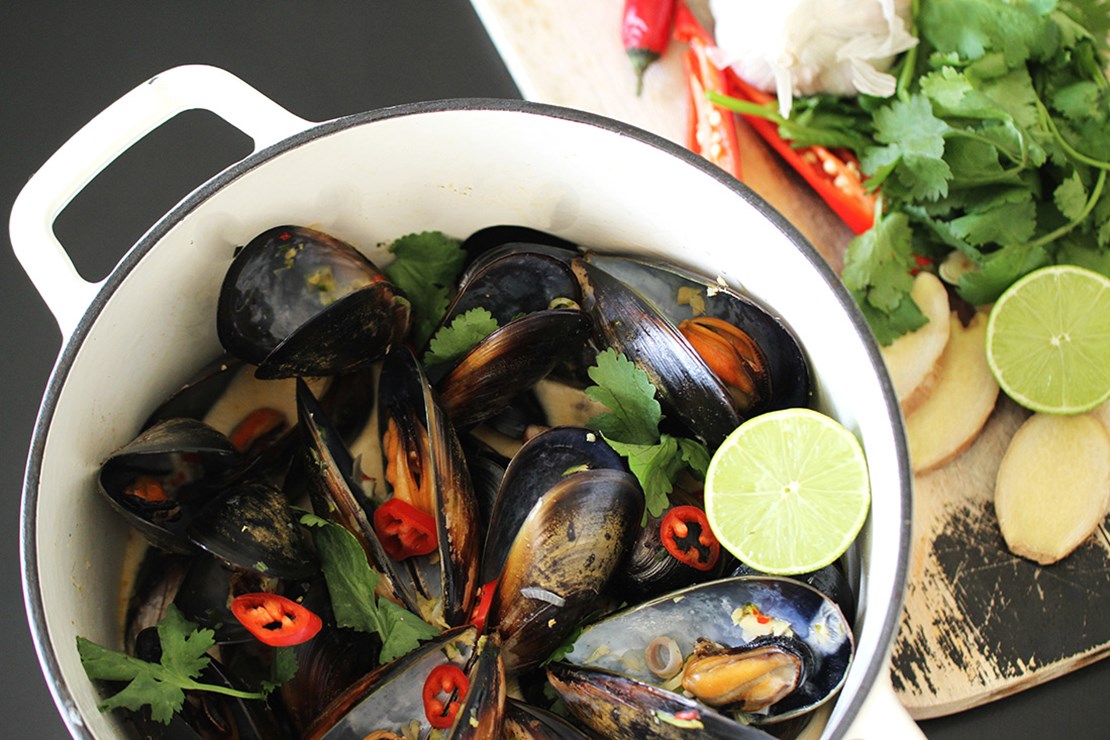 A pot of fresh mussels garnished with slices of red chili, cilantro, and lime wedges, ready for cooking or serving. The mussels are arranged in a white pot, and additional garnishes are placed on a wooden cutting board next to it.