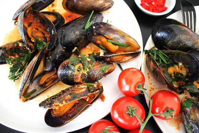 A plate of cooked mussels garnished with herbs, accompanied by fresh cherry tomatoes, presented on a white plate.