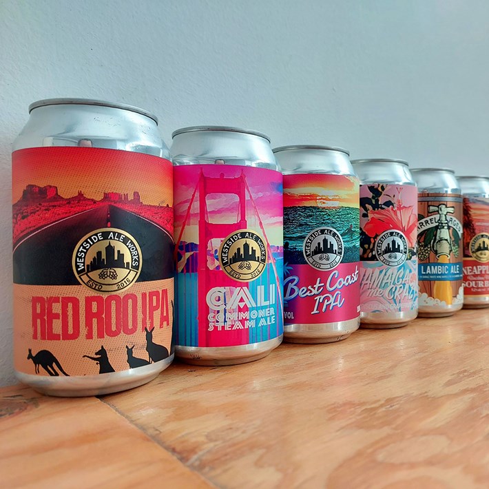 A row of various colorful beer cans from Westside Ale Project with artistic labels, each depicting a different flavor, lined up on a wooden surface against a light blue wall.
