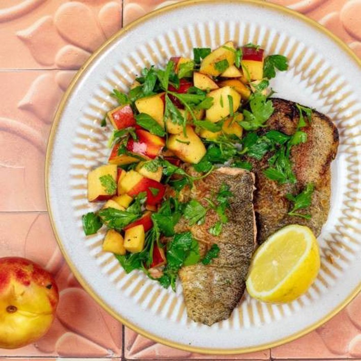 A round ceramic plate with a golden rim holds two crispy, golden-brown fish fillets on the right and a vibrant salad on the left. The salad consists of diced nectarines with red skin, fresh green parsley, and herbs. A bright yellow lemon wedge sits near the fish. The plate rests on a pinkish-orange tiled surface with a floral pattern, and a whole nectarine is placed nearby.