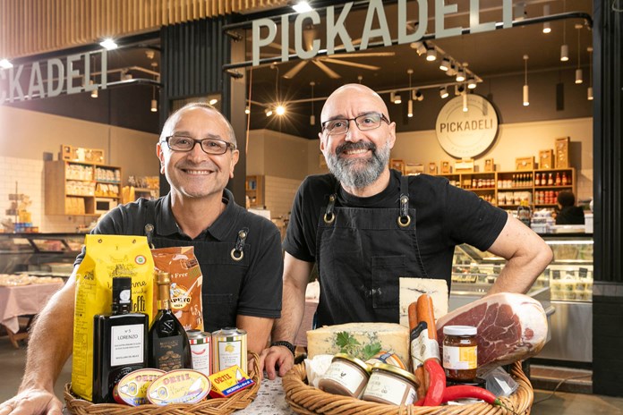 Two individuals standing in front of a store named ‘PICKADELI’, holding baskets filled with various gourmet food items.