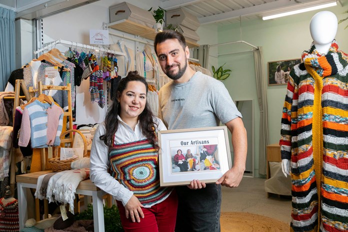 Two people holding a framed picture titled ‘Our Artisans’, standing in a well-lit store with various clothing items displayed around them.