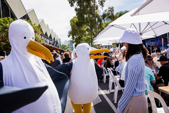 A vibrant outdoor event featuring people interacting with large, whimsical seagull puppets under a bright sky. Two seagull puppets with white bodies, black wings, and orange beaks take center stage. Attendees, including one person in a white hat and grey sweater, engage with the puppets. White chairs, tables, and umbrellas dot the area, creating a relaxed atmosphere. In the background, a crowd gathers near tents and buildings. 