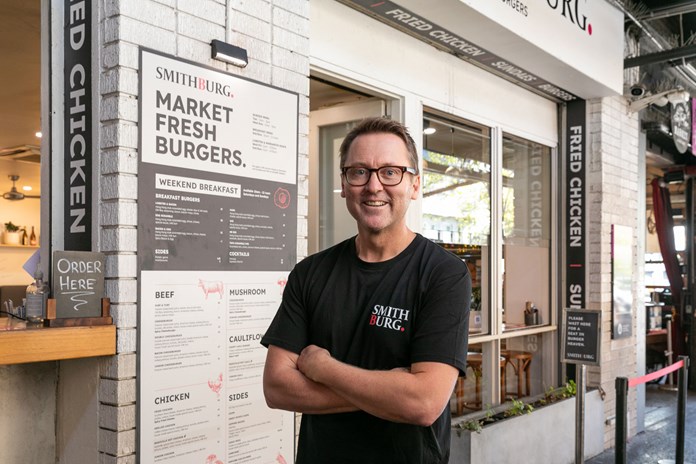 The image shows an individual in a black t-shirt with “SMITH BURG” printed on it, standing in front of a menu board titled “SMITHBURG MARKET FRESH BURGERS.” The menu lists various food options, including weekend breakfast and different types of burgers and sides. An “ORDER HERE” sign is to the left of the menu, and signs for “FRIED CHICKEN” and “BURGERS” are visible in the background. The setting appears to be a modern, well-lit fast-casual restaurant.