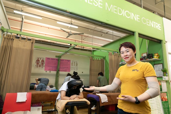 The image shows a person in a yellow shirt standing in front of Dr Shen's Chinese Medicine . Other individuals are receiving treatments on wooden beds with beige curtains. The centre’s signboard is green with white text, and various informational posters adorn the walls. The person in the foreground appears to be wearing a brace on their left arm.