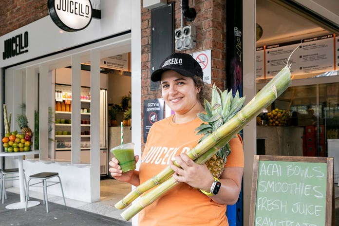 Person in an orange t-shirt, holding a green smoothie and a large stalk of sugar cane with a pineapple attached, standing in front of ‘JUICELO’, a juice bar.