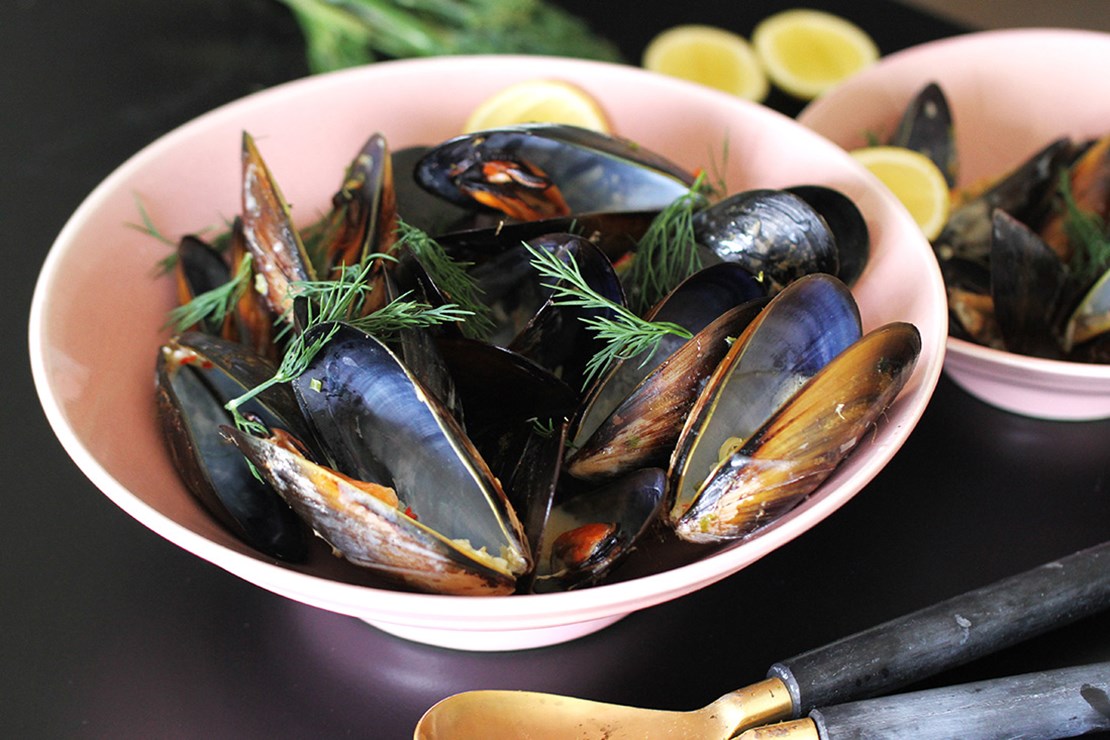 A bowl of fresh mussels garnished with dill and lemon wedges, ready to be served. The mussels are dark-shelled, glossy, and opened up, revealing the cooked insides. Lemon wedges add a pop of color in the background. An antique-looking metal spoon with a golden handle rests on the surface to the right.