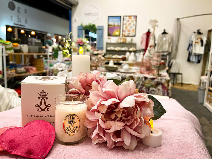The image features a charming boutique store interior with a pink table at the forefront displaying a white box labeled “CHATEAU DE VERSAILLES”, a lit candle, and a bouquet of pink flowers, creating a warm and inviting atmosphere. The background is filled with various items for sale, including clothing and decorative pieces, all bathed in soft lighting.