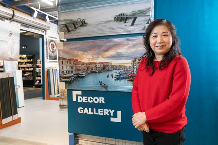 Person in a red sweater standing next to a blue wall displaying various framed pictures, including a prominent one of a scenic waterfront cityscape, inside a store labeled ‘DECOR GALLERY